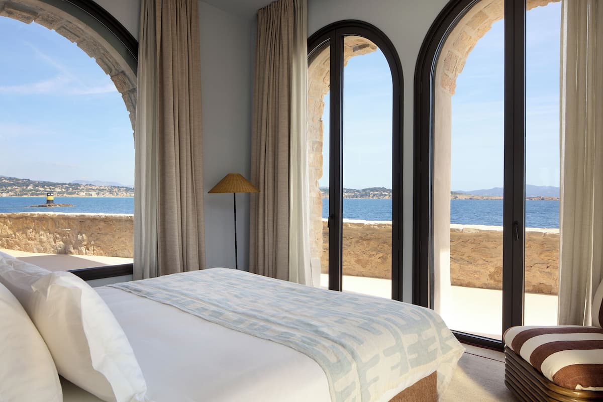 A bedroom with a large bed, a lamp, and a view of the sea through arched windows.