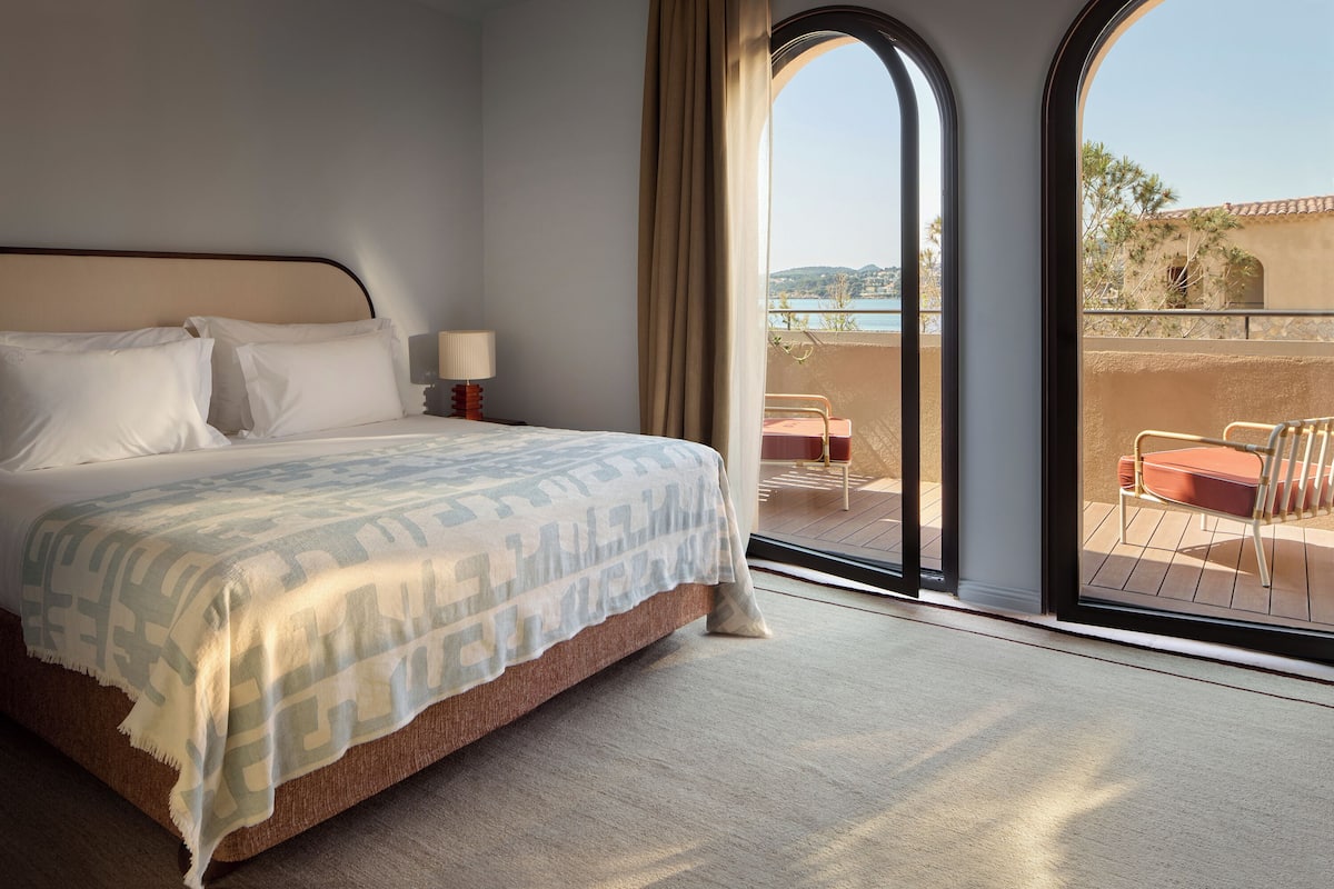 A hotel room with a bed, a nightstand, a lamp, and a balcony with a view of the sea.