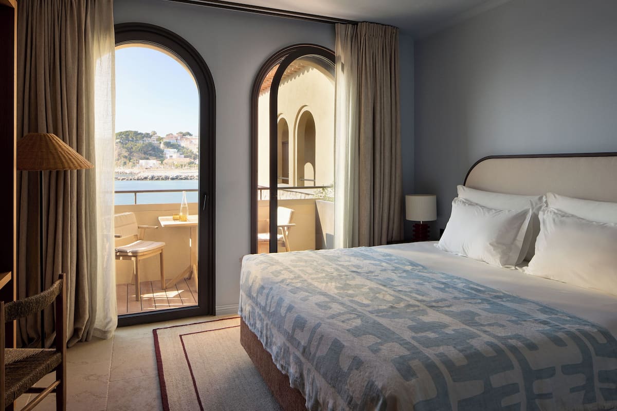 A hotel room with a bed, a chair, a table, and a view of the sea.