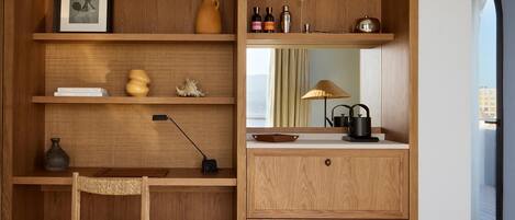 Escritorio de madera con una silla, estanterías con libros y objetos decorativos, un espejo y una zona de cocina con hervidor y tetera.