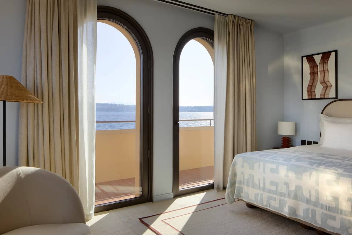 A hotel room with a bed, a chair, a lamp, a nightstand, and a painting. There are two large arched windows with curtains, and a view of the sea.