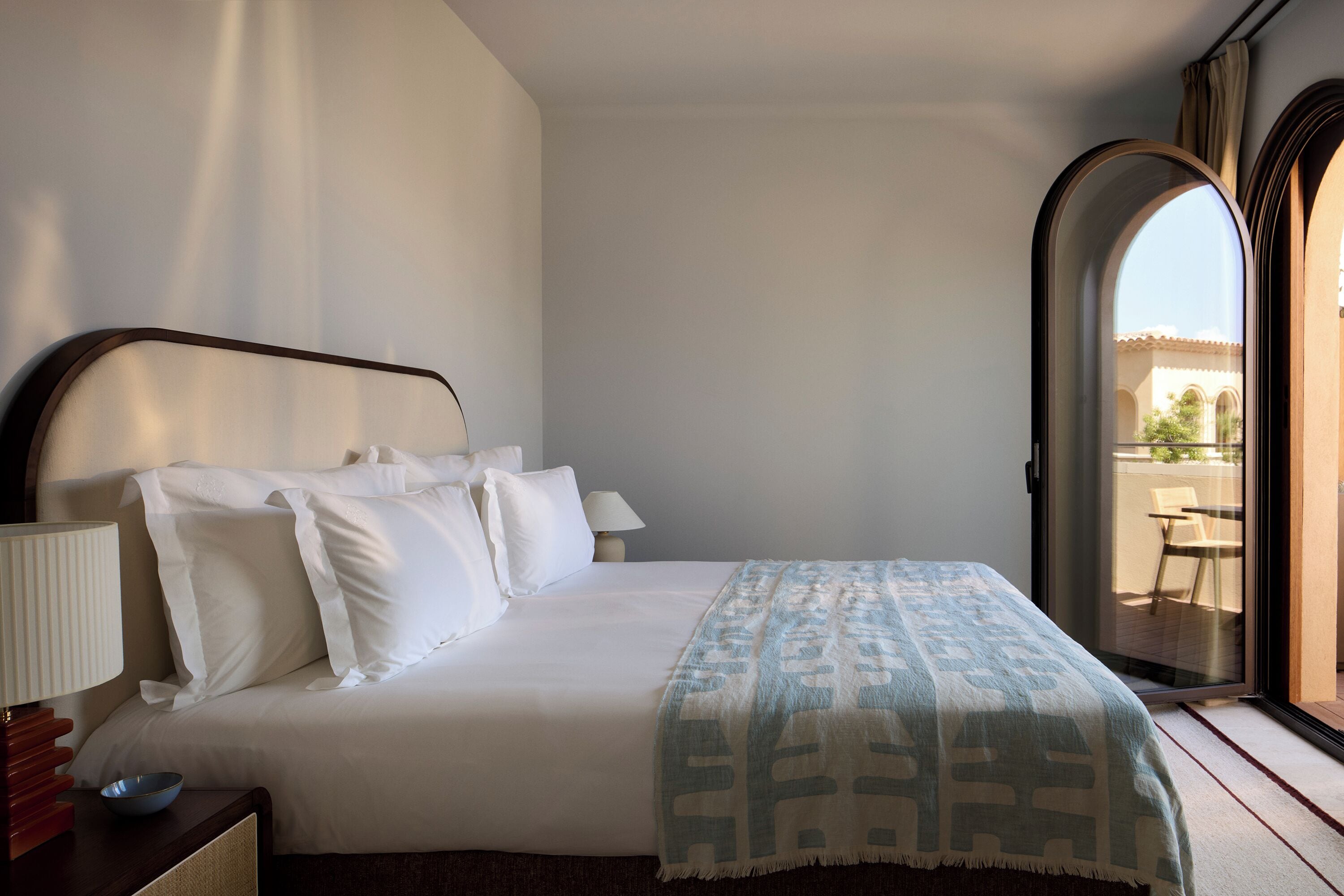 Quarto de hotel com cama, mesa de cabeceira, abajur e vista para o exterior por uma porta em arco.