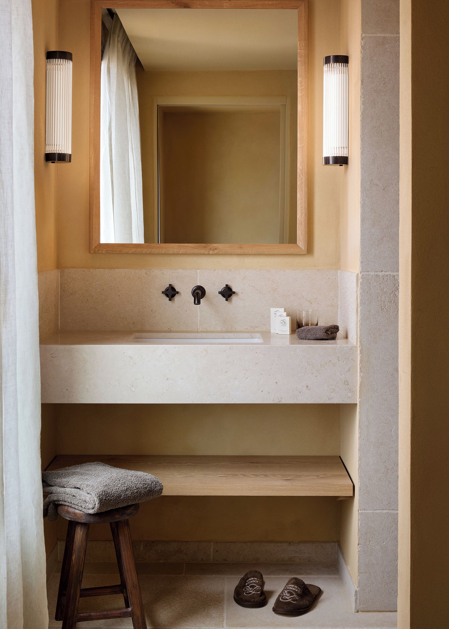 A bathroom with a wooden stool, a sink with three knobs, and a mirror above it.