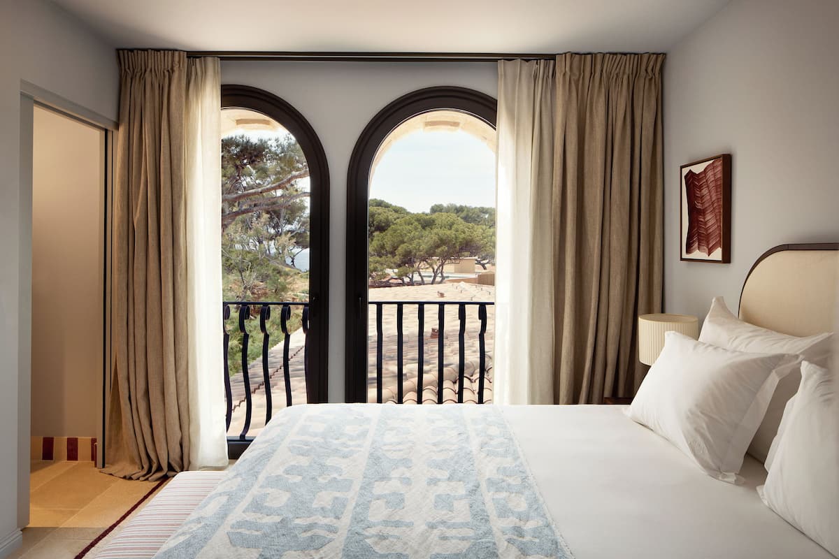 A bedroom with a large bed, two windows with curtains, a view of trees and a balcony, and a framed picture on the wall.