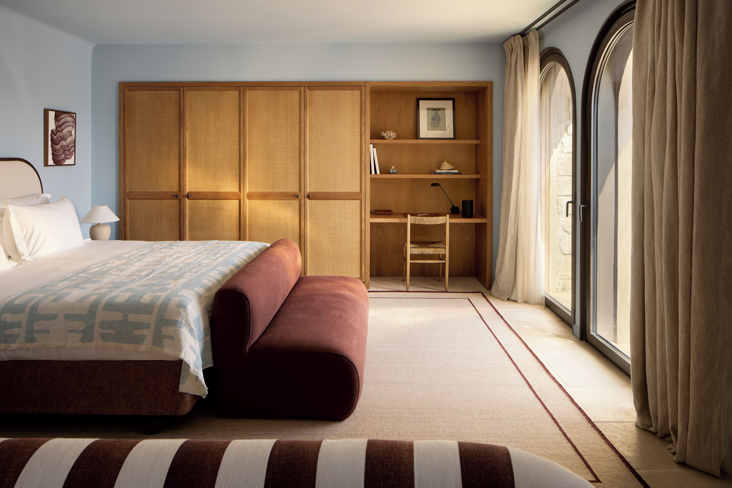 A bedroom with a bed, a wooden wardrobe, a chair, and a large window with curtains.