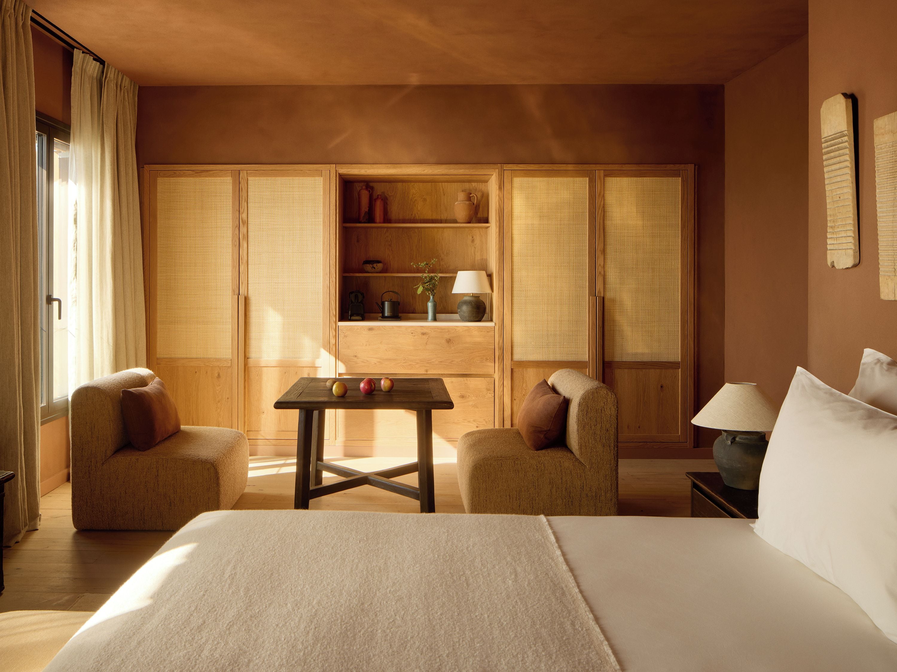 Habitación de hotel con una cama, dos sillones, una mesita con manzanas, una lámpara y una estantería decorativa.