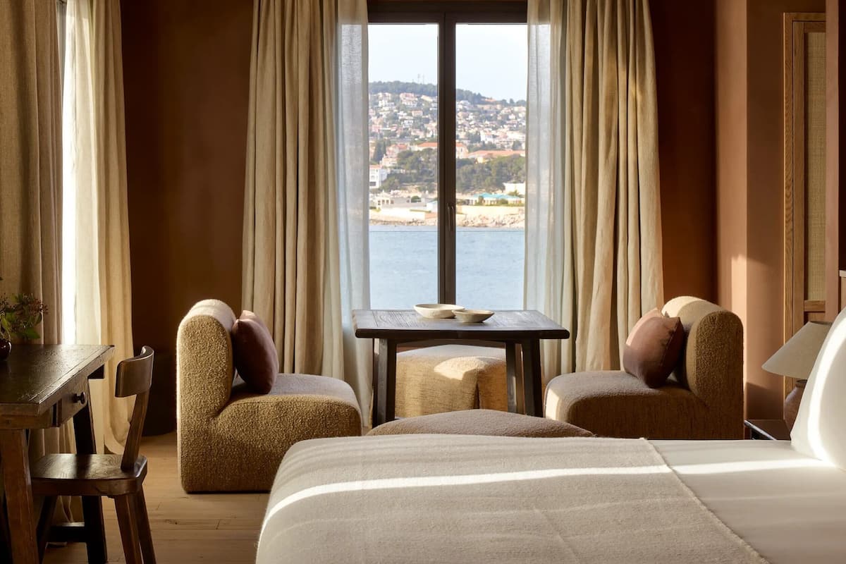 A hotel room with a bed, two armchairs, a small table with a bowl, and a view of a coastal town through large windows.
