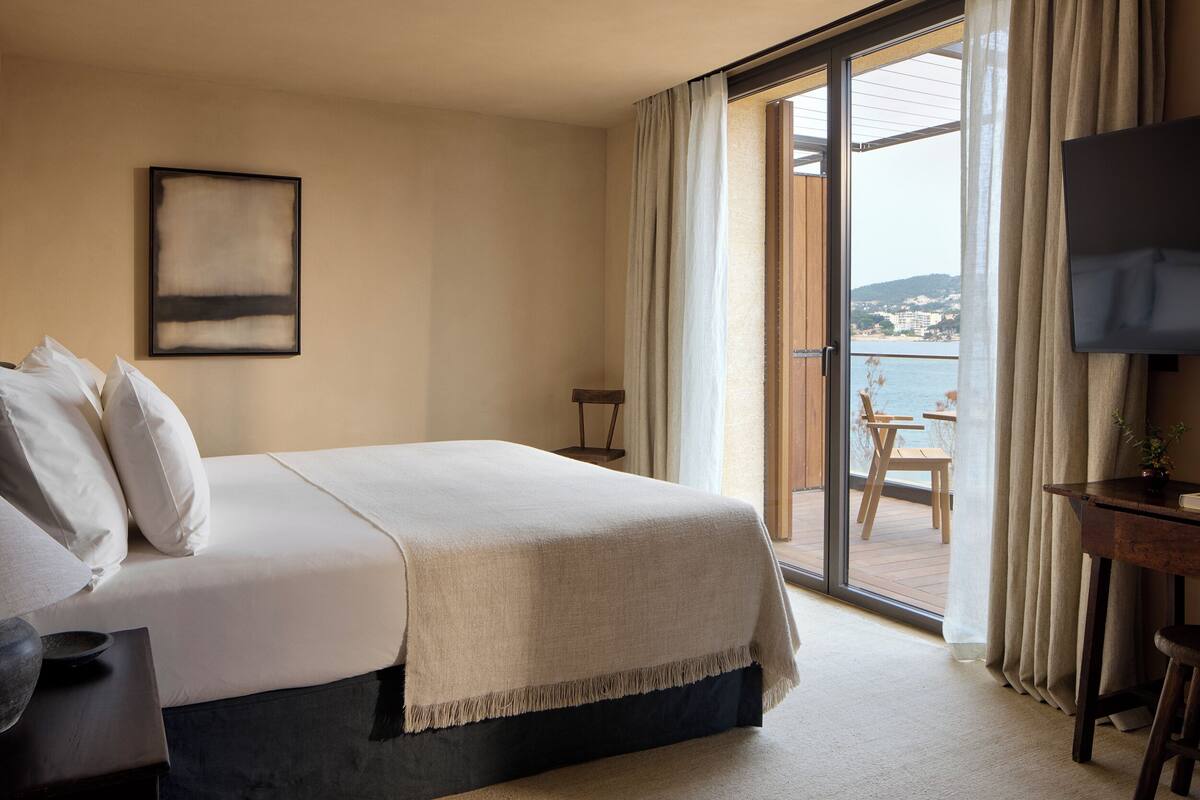 A hotel room with a bed, a TV, a balcony with a table and chairs, and a view of the sea.