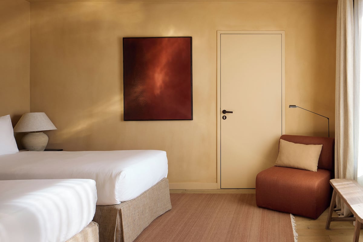 A hotel room with two beds, a desk, a chair, a lamp, a door, and a painting on the wall.