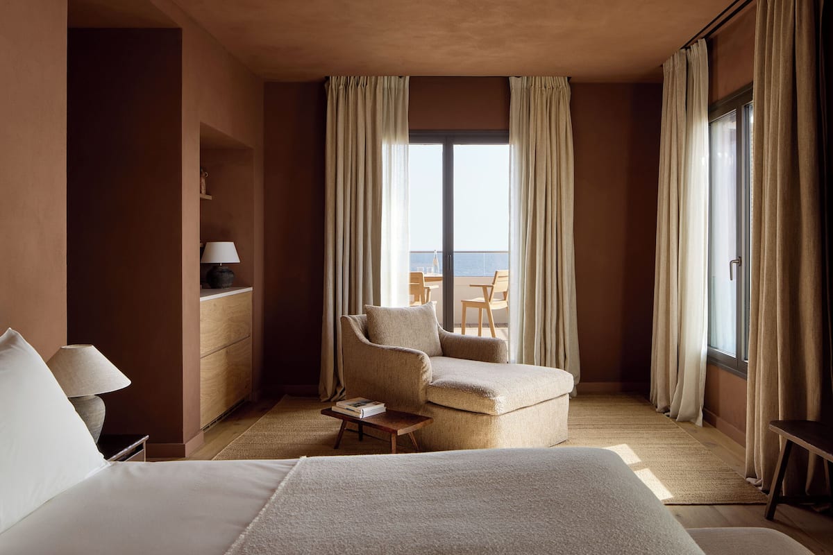 A hotel room with a bed, a chair, a small table, a lamp, and a view of the sea through large windows.