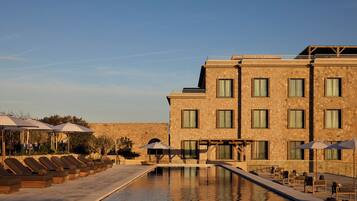 Un área de piscina con sillones de descanso y sombrillas, un edificio de piedra con múltiples ventanas, y un cielo despejado.