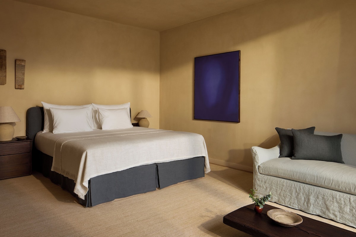 A bedroom with a bed, bedside tables, a sofa, a wall-mounted painting, and a small plant on a side table.