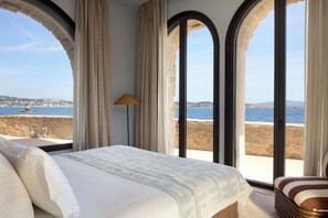 Ein Schlafzimmer mit einem großen Bett, einer Lampe und Meerblick durch bogenförmige Fenster.