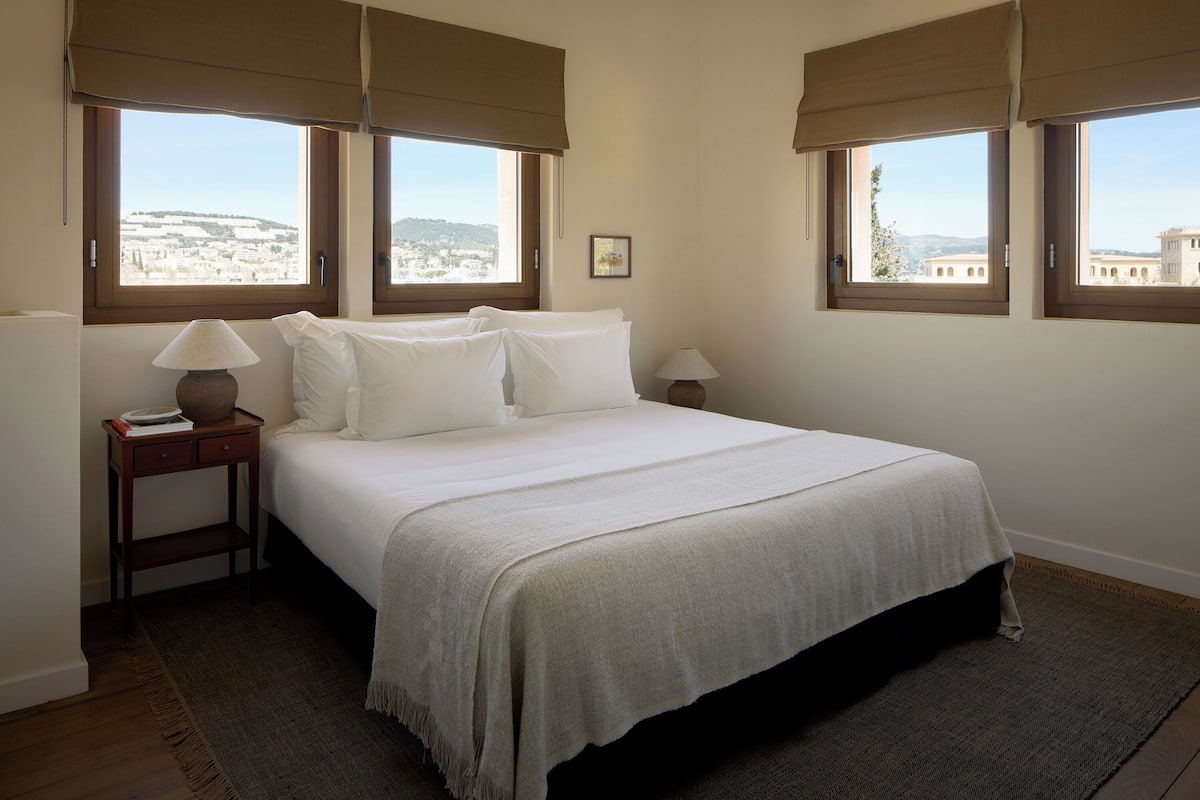 A bedroom with a bed, two bedside tables, a lamp, and a view of a cityscape through large windows.