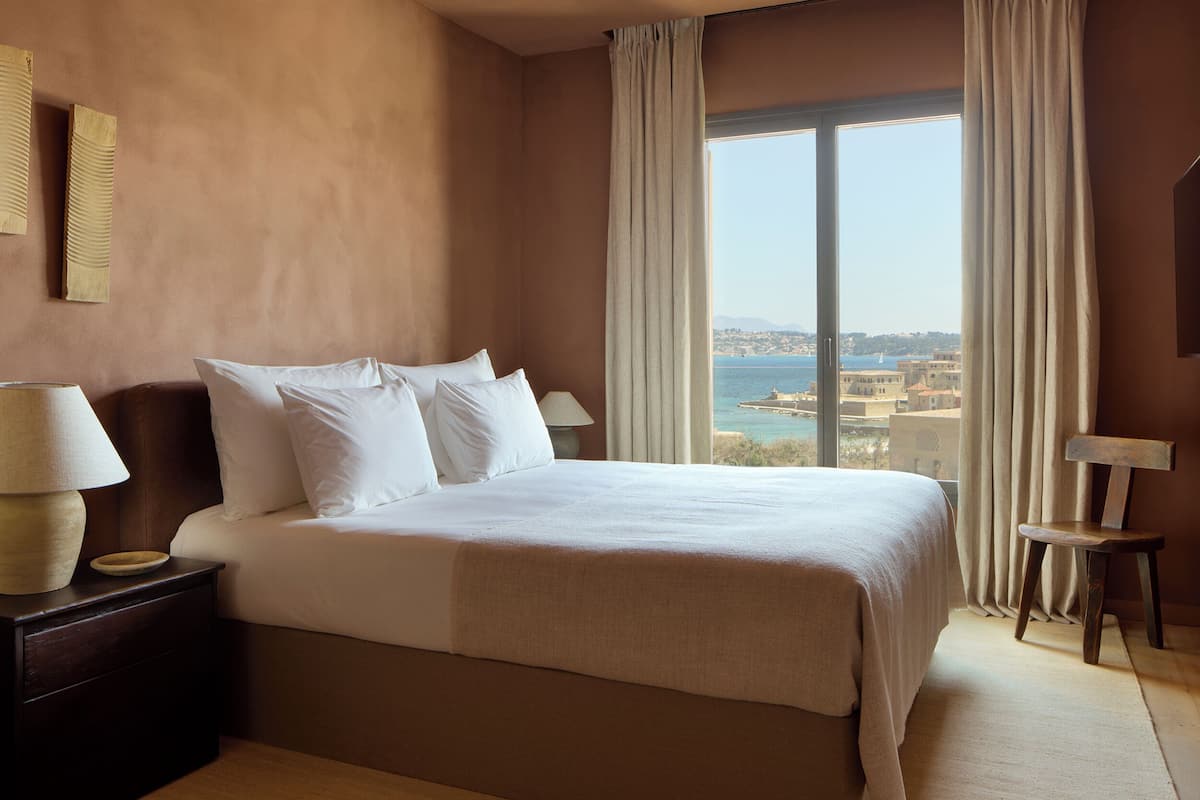 A hotel room with a large bed, a nightstand with a lamp, a chair, and a view of the sea through a large window.