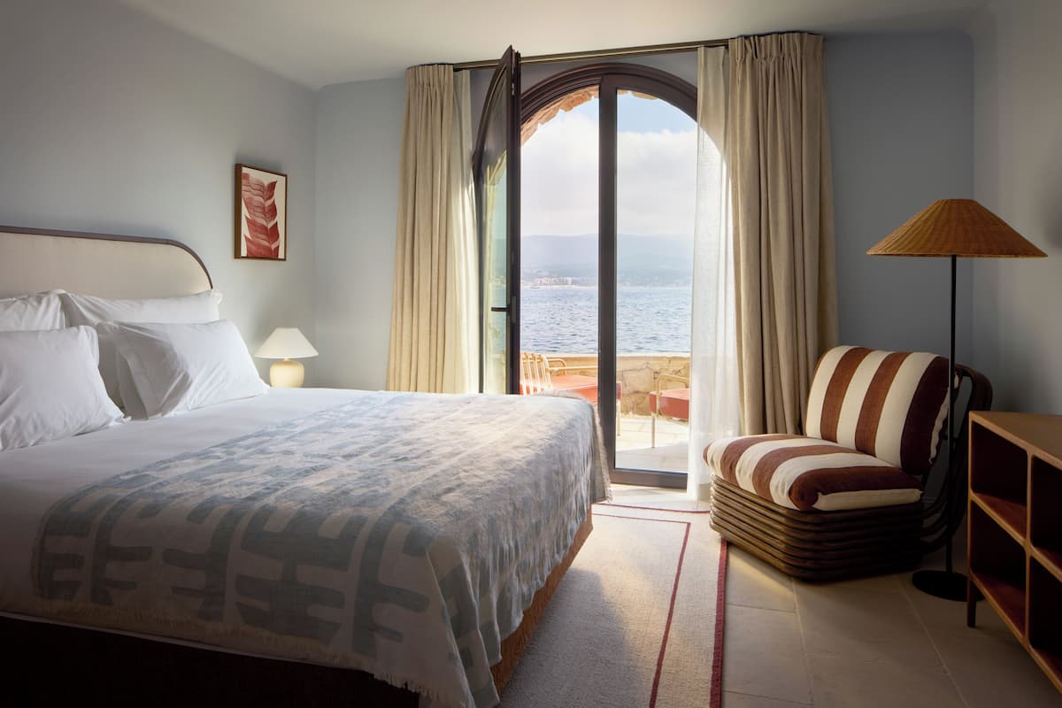 A hotel room with a bed, a chair, a lamp, a view of the sea, and a painting on the wall.