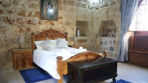Una camera da letto con parete in pietra, un letto in legno con poggiatesta, un comodino, una panca e una finestra con tende.