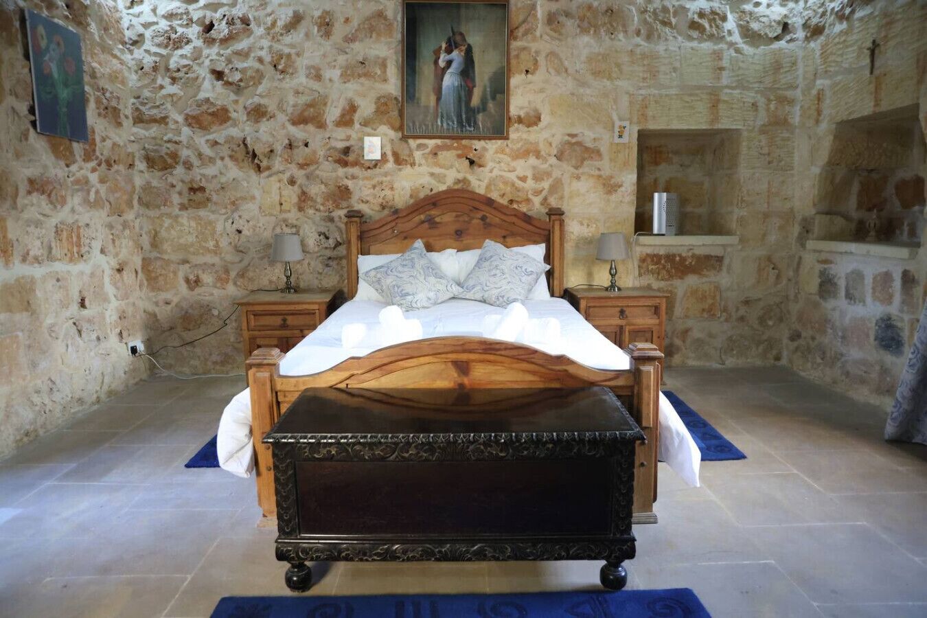 A bedroom with a stone wall, a wooden bed, a nightstand, and a painting on the wall.