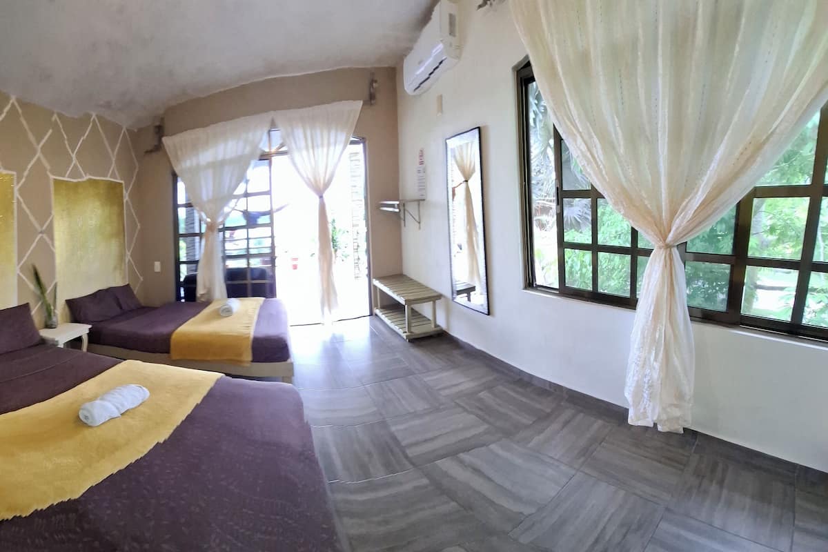Una habitación de hotel con dos camas, un ventanal con cortinas, un aire acondicionado instalado en la pared y una mesita pequeña.