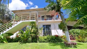 Ein zweigeschossiges Gebäude mit einer Treppe, die zu einem Balkon führt, umgeben von üppiger tropischer Vegetation.