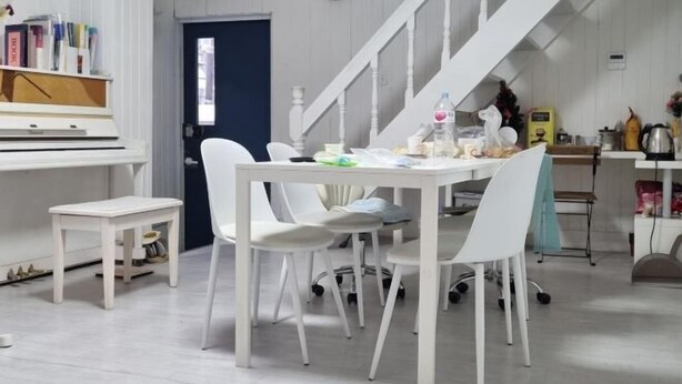 A room with a white piano, a dining table with chairs, a staircase, and a kitchenette area.