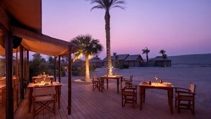An outdoor dining area with tables and chairs, palm trees, and a view of the beach and ocean at dusk. - The Outpost Al Barari Camp (Al Wakrah)