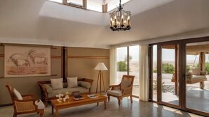 A spacious living room with a sofa, chairs, a coffee table, and a chandelier. There is a large window with a view of the outdoors. - The Outpost Al Barari Camp (Al Wakrah)