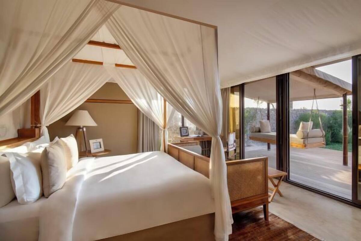 A bedroom with a canopy bed, a bedside table with a lamp, and a view of an outdoor seating area.