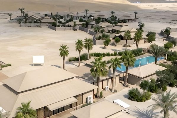 An aerial view of a resort with multiple buildings, palm trees, and a swimming pool. - The Outpost Al Barari Camp (Al Wakrah)