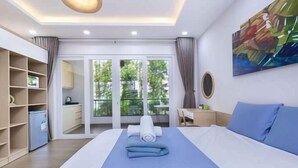 A hotel room with a bed, a desk, a chair, a wardrobe, and a balcony with a view of greenery. - Cozrum Homes - Yoga Corner (Ho Chi Minh City)
