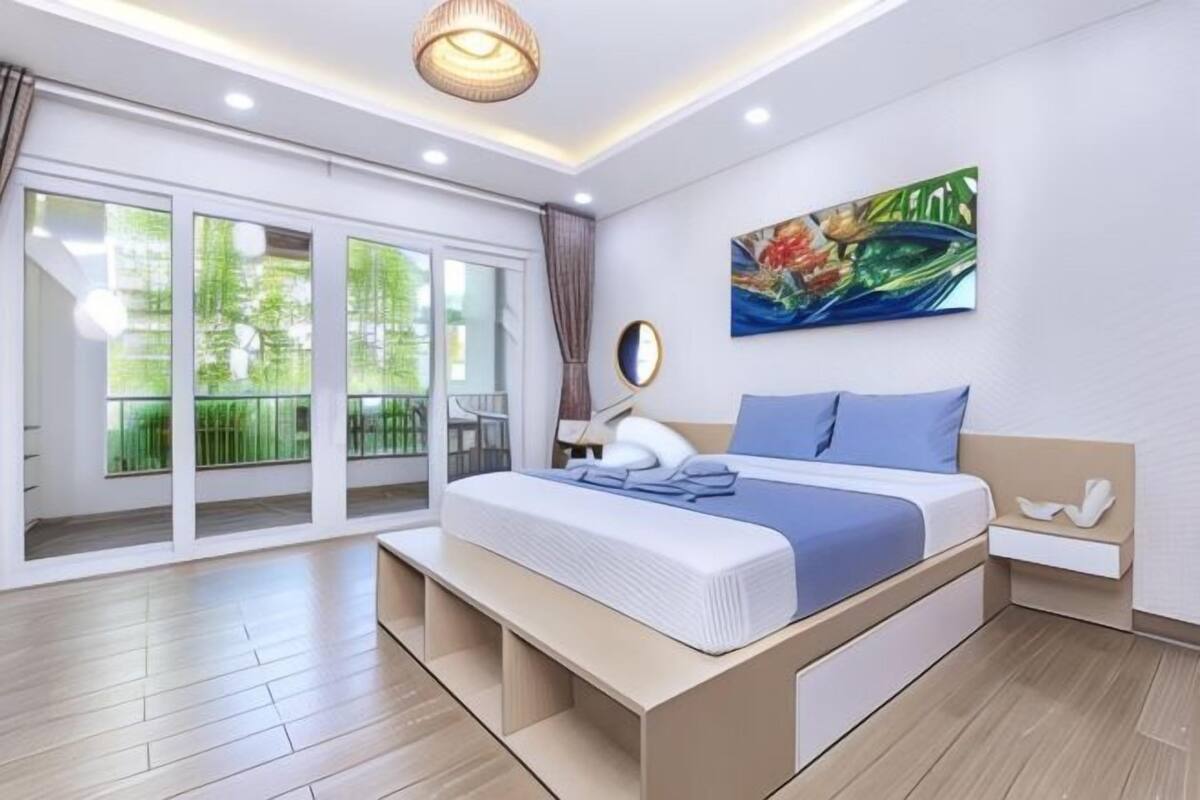 Une chambre moderne avec un grand lit, une table de chevet, une œuvre d’art accrochée au mur et une porte-fenêtre coulissante donnant sur un balcon.