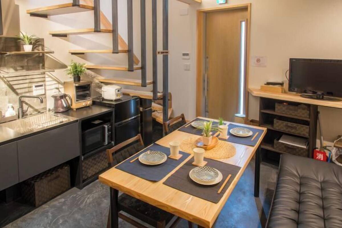Une cuisine moderne avec une table en bois dressée pour le repas, un escalier et un téléviseur posé sur une étagère.