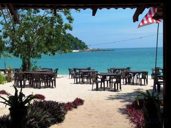 An outdoor seating area with tables and chairs facing a beach, under a covered structure with an American flag. - Forget-Me-Not Pangkor Homestay 70 (Pangkor)