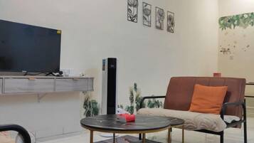 A room with a flat-screen TV, a sofa with an orange cushion, a small round table with a red object, and a wall-mounted cabinet.