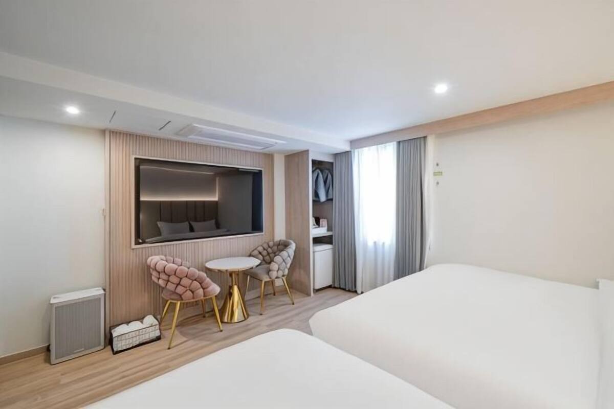 A modern hotel room with a bed, a small table, two chairs, a TV, and a window with curtains.