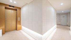 A modern hallway with a wooden elevator, a white wall with a recessed panel, and a light-colored floor. - Masan Den Basta hotel Gyeongnam Uni (Changwon)