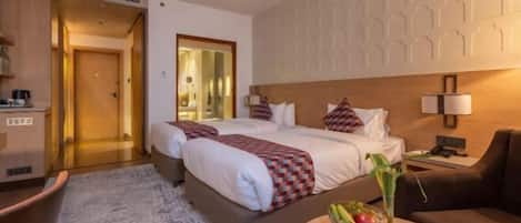 A hotel room with two beds, a sitting area, a small table with a vase, and a view of the hallway.
