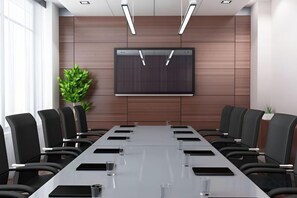 Un salón de conferencias con una larga mesa, sillas, una pantalla de proyector y una planta en maceta.