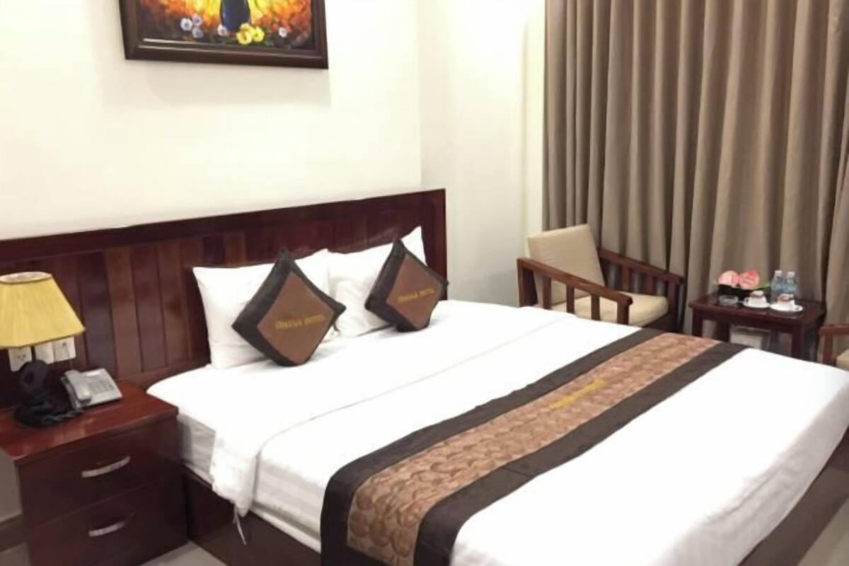 Une chambre d’hôtel avec un lit, une table de chevet avec une lampe, une chaise, une petite table avec un téléphone et un vase, et un rideau.