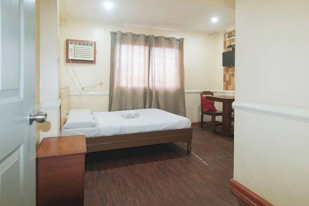 A small, neatly arranged bedroom with a single bed, a bedside table, a chair, and a small table with a red chair.