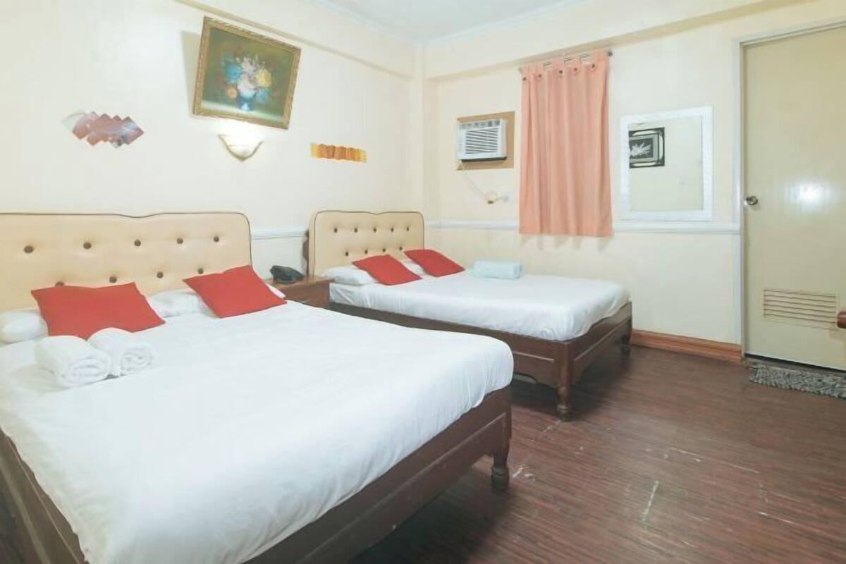 A hotel room with two beds, each with white linens and red pillows. There is a painting on the wall, a window with a curtain, and an air conditioning unit.