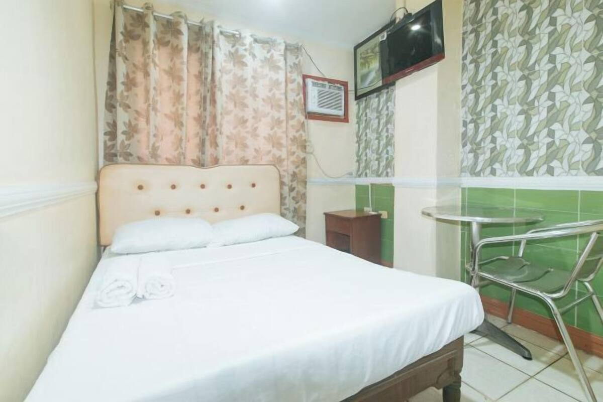 A single bed with a wooden headboard, a small bedside table, a TV mounted on the wall, and a green-tiled wall with a patterned design.
