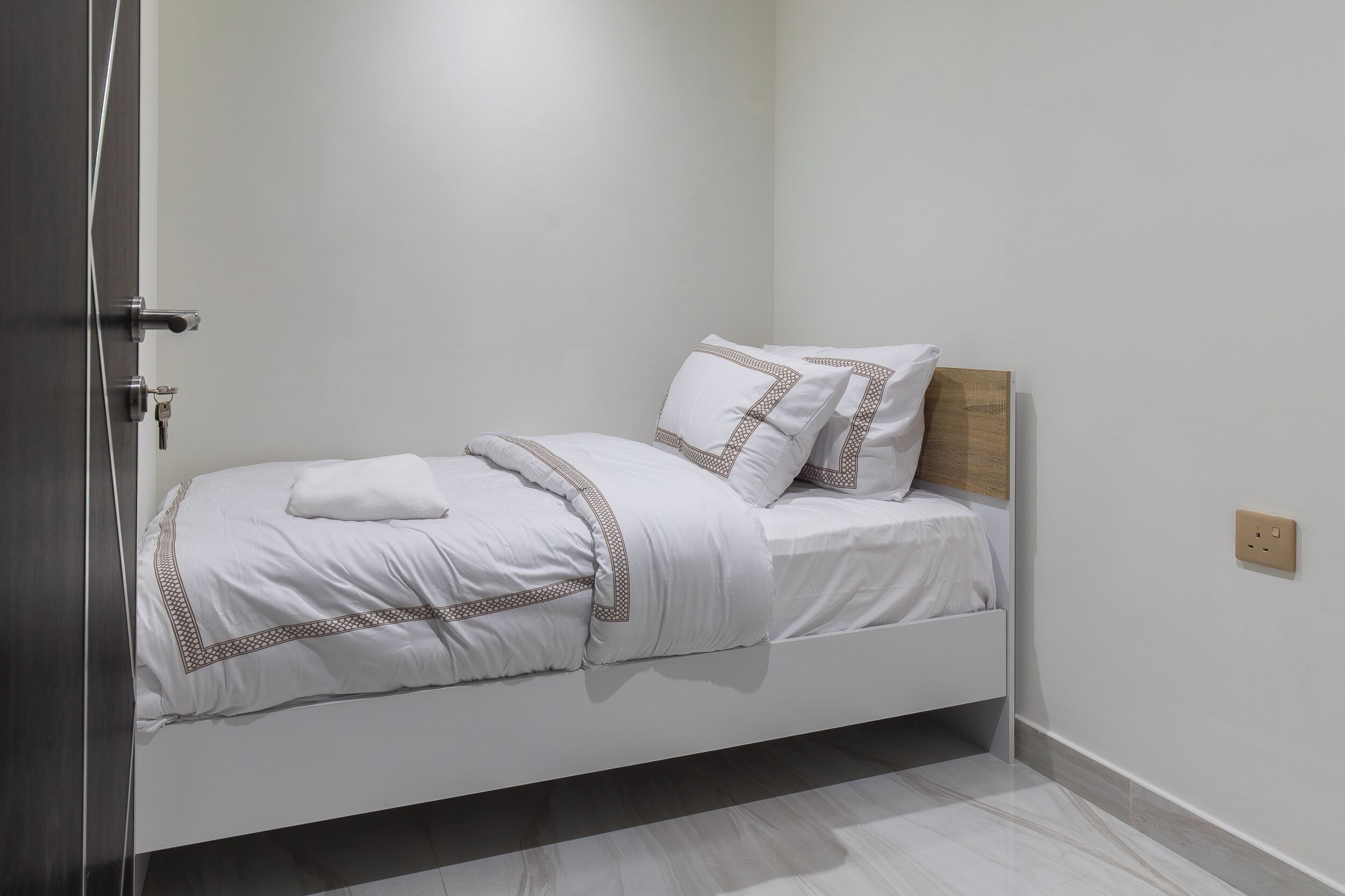 A single bed with white bedding and a wooden headboard in a room with a plain white wall and a door on the left.