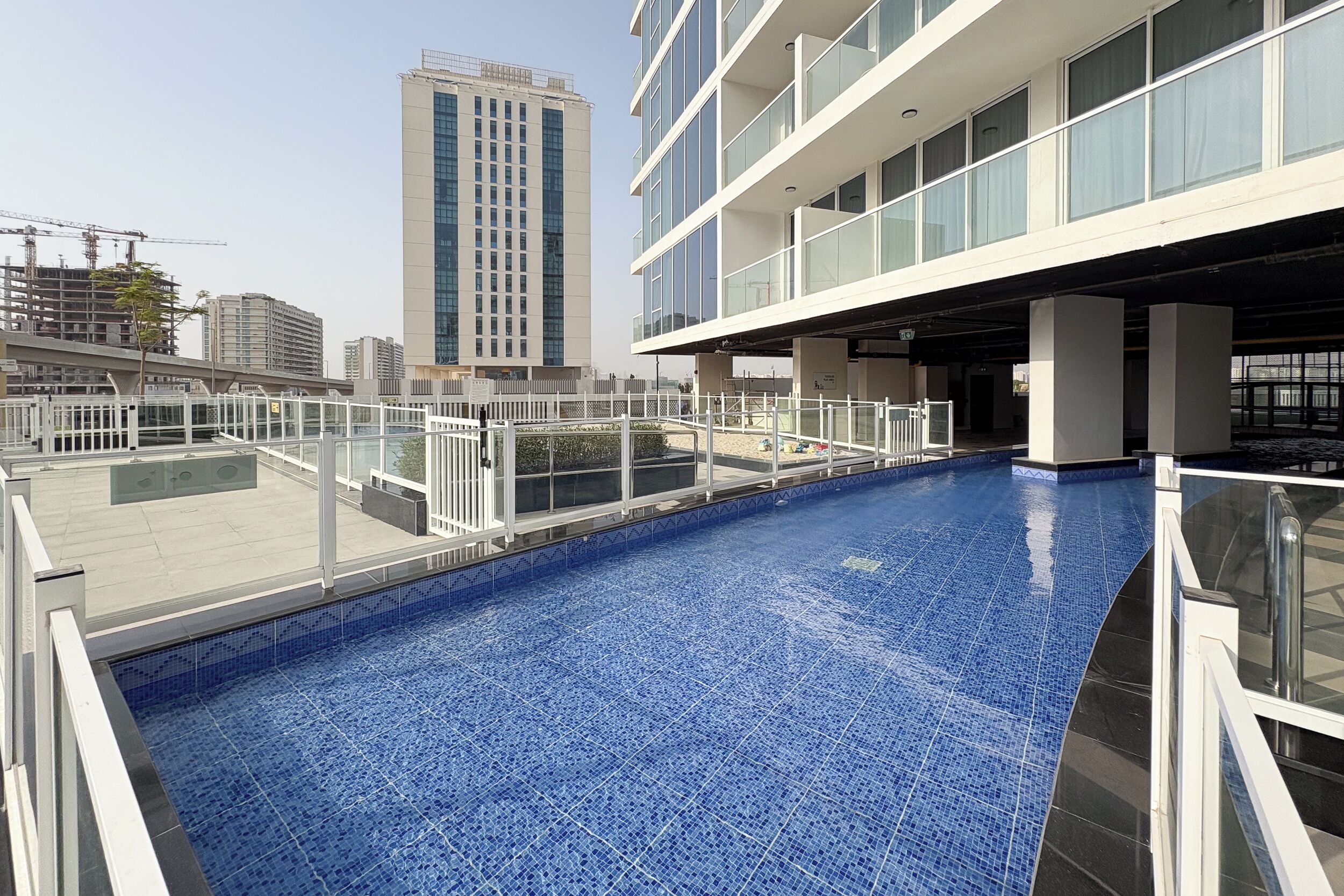 A rooftop swimming pool with a blue tiled floor, surrounded by a white railing, and adjacent to a modern building with large windows.