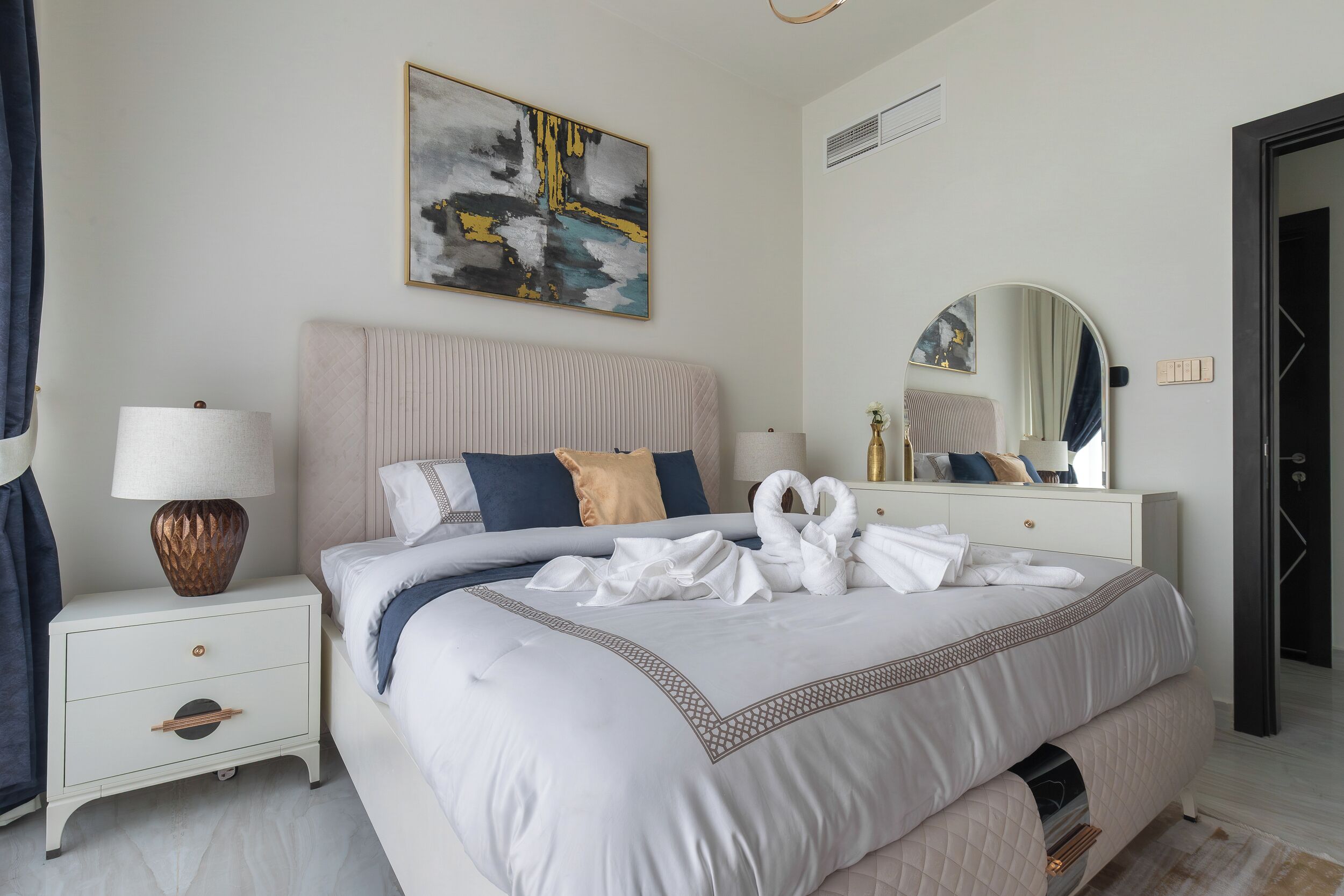 Una cama bien hecha con una decoración de almohadones en forma de cisne, una lámpara de noche y un cuadro abstracto colgado en la pared.