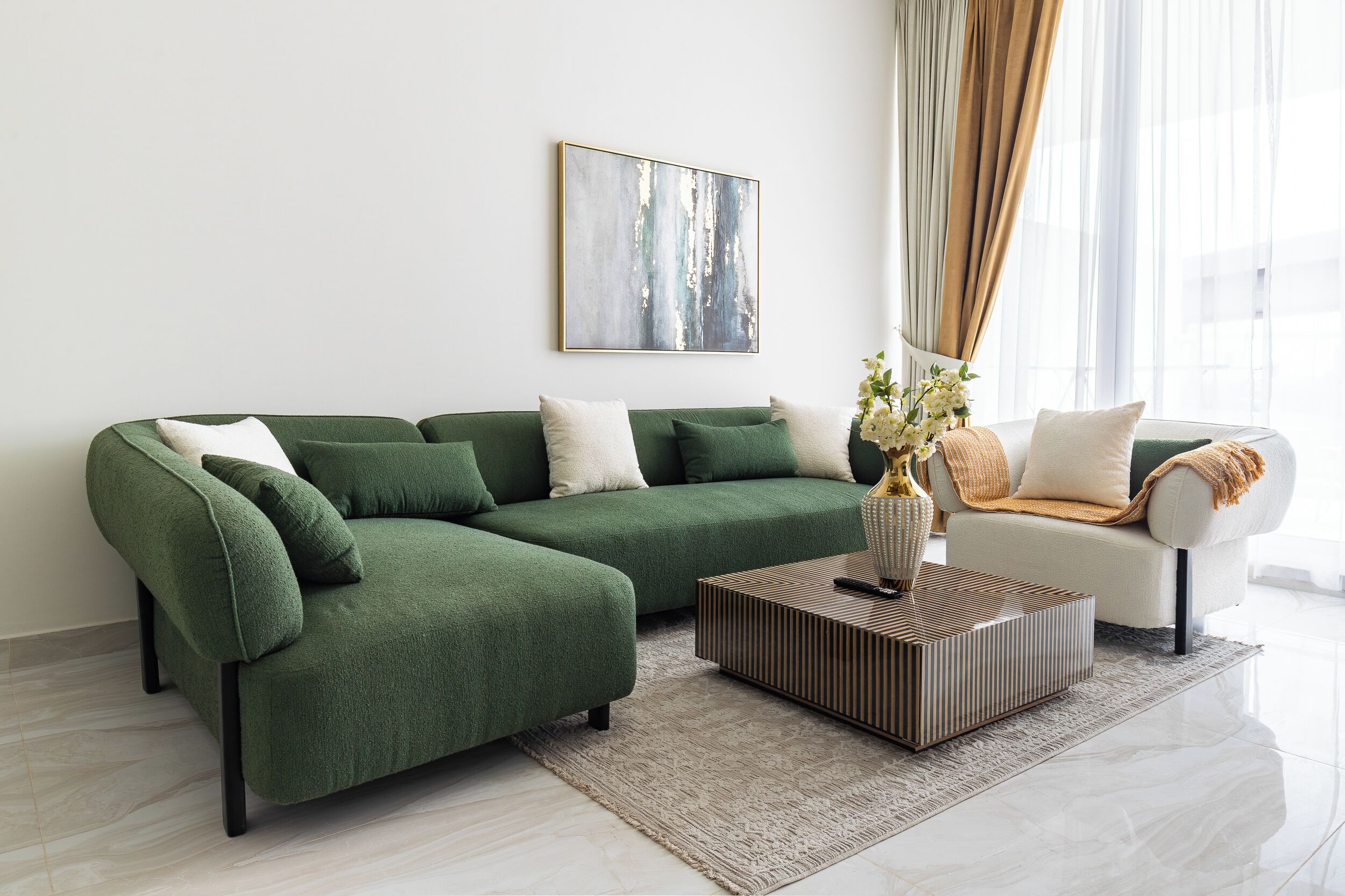 Una sala de estar con un sofá seccional verde, un sillón blanco, una mesa de centro y un cuadro en la pared.
