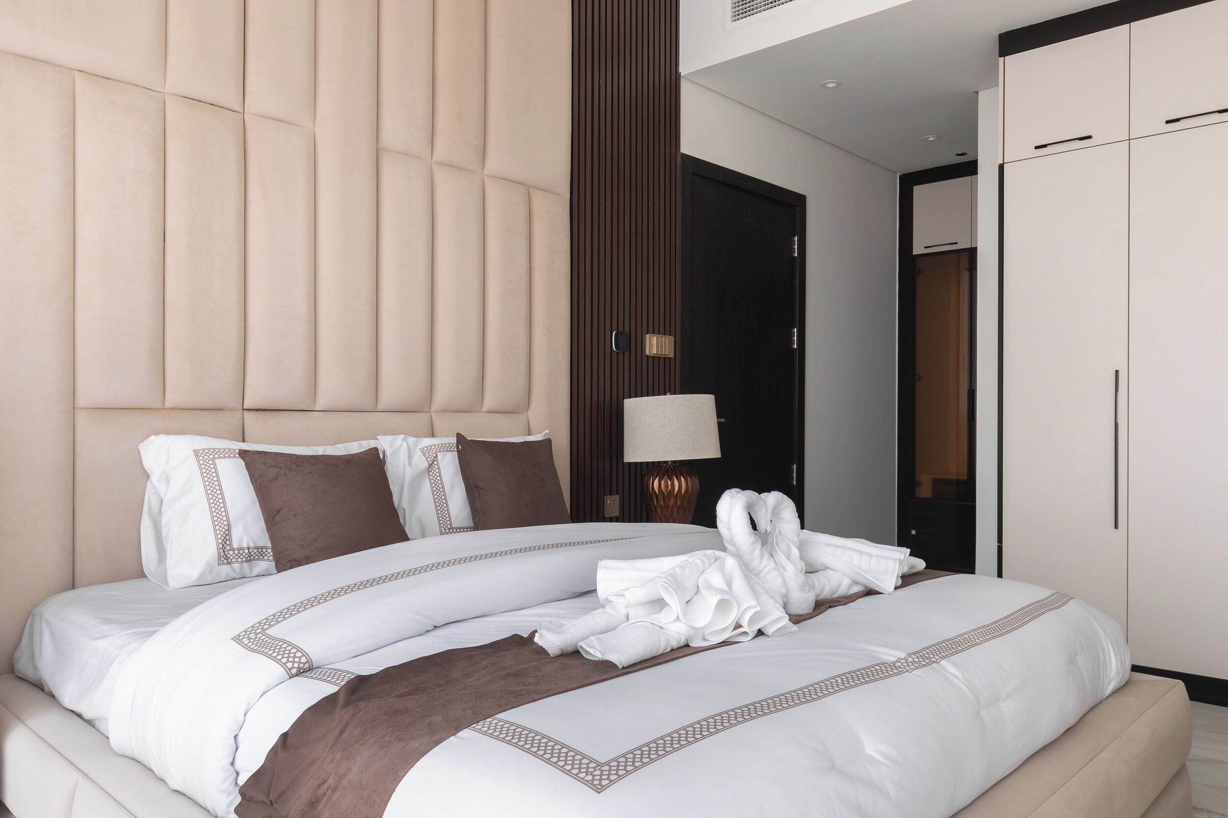 A modern hotel room with a large bed, a headboard, a wardrobe, and a nightstand with a lamp.