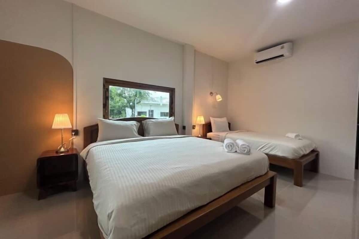 A hotel room with a large bed, two bedside tables with lamps, a window with a view of greenery, and an air conditioning unit on the wall.