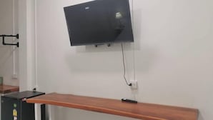Un téléviseur fixé au mur sur un mur blanc, un banc en bois et un petit objet noir posé sur le banc.