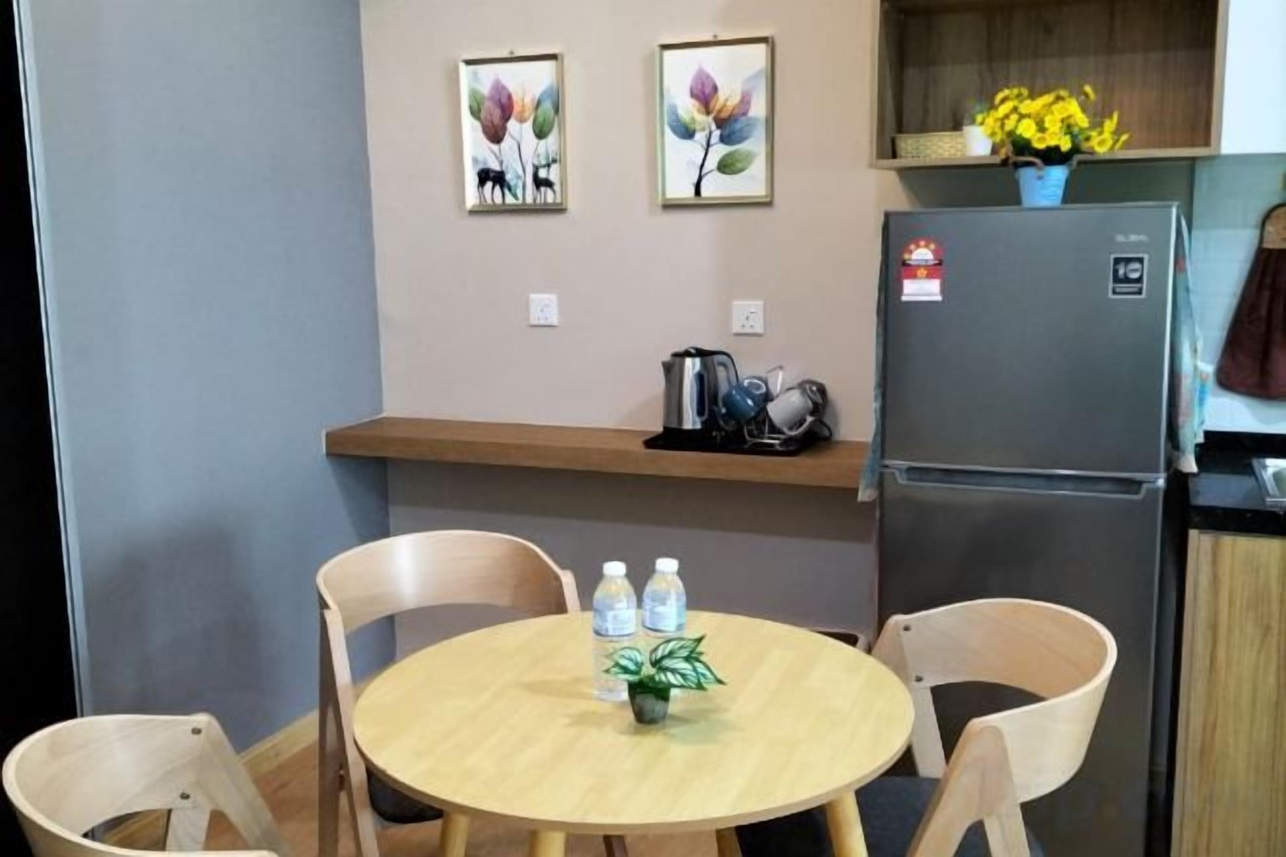 A small dining area with a round table, two chairs, a refrigerator, and a shelf with flowers.